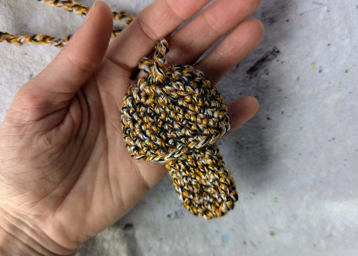Ein gehäkelter Feuerzeughalter zum Umhängen in Pilzoptik in gelb, schwarz und weiß gemischten Farben liegt in einer Hand vor weißem Hintergrund.