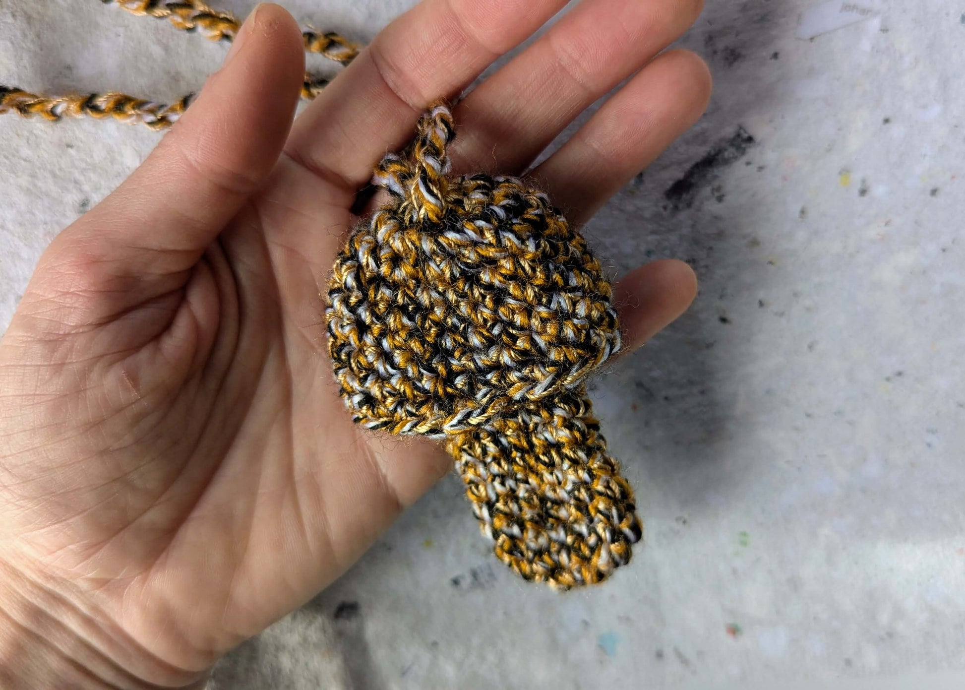 Ein gehäkelter Feuerzeughalter zum Umhängen in Pilzoptik in gelb, schwarz und weiß gemischten Farben liegt in einer Hand vor weißem Hintergrund.