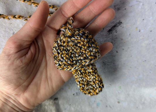 Ein gehäkelter Feuerzeughalter zum Umhängen in Pilzoptik in gelb, schwarz und weiß gemischten Farben liegt in einer Hand vor weißem Hintergrund.