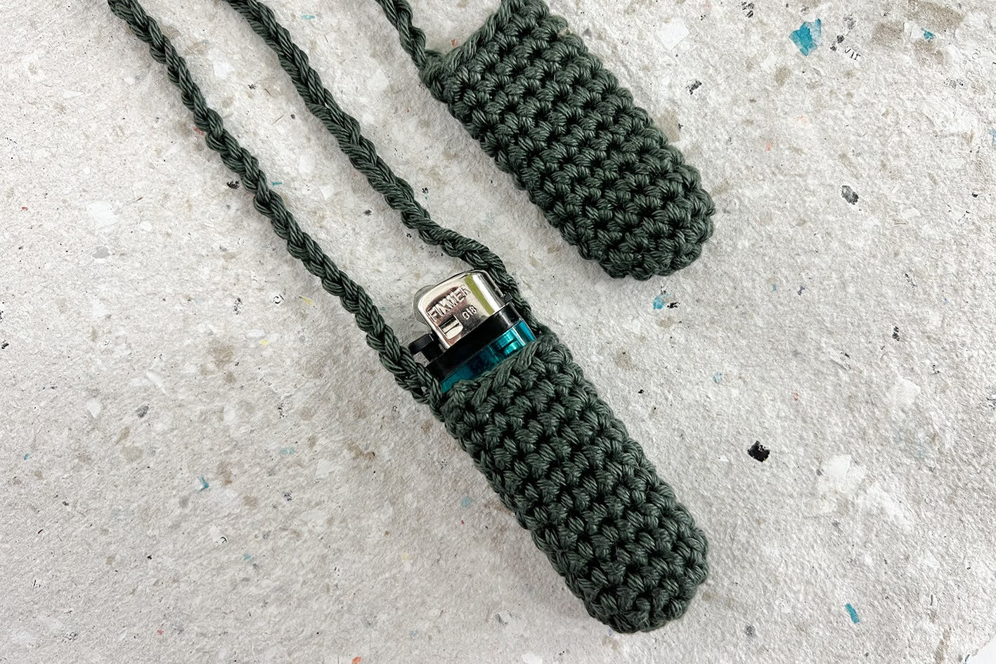 Zwei gehäkelte Taschen fürs Feuerzeug zum Umhängen in olivgrün. Eine Tasche trägt ein blaues Feuerzeug, die andere Tasche ist leer.