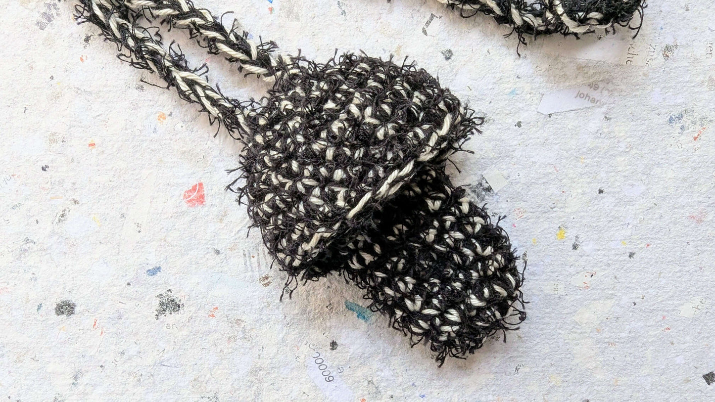 Ein gehäkelter Feuerzeughalter zum Umhängen in Pilzoptik in schwarz und weiß  liegt auf weißem Hintergrund.