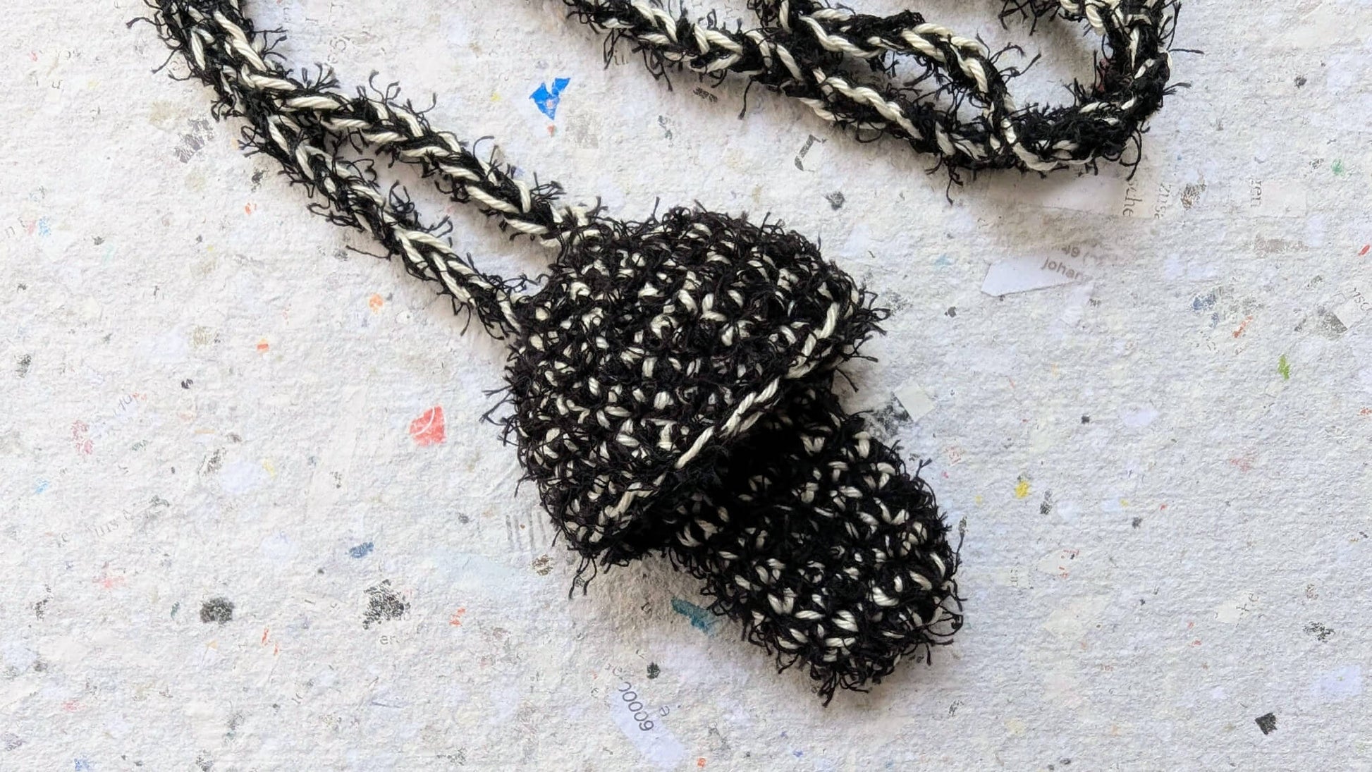 Ein gehäkelter Feuerzeughalter zum Umhängen in Pilzoptik in schwarz und weiß  liegt auf weißem Hintergrund.