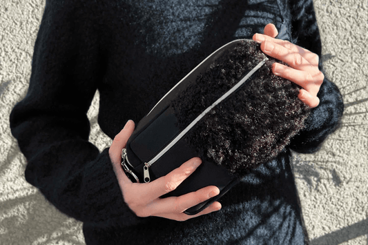 Frau hält schwarze Bauchtasche aus Upclycling-Materialien mit silbernem Reißverschluss in beiden Händen. Die Bauchtasche besteht teilweise aus einem flauschigen Material.