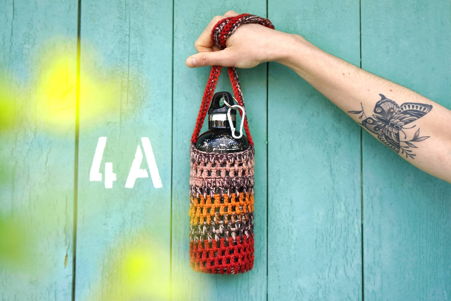 Gehäkelter rot brauner Flaschenhalter CHRISTA von jeff von Frauenhand ins Bild gehalten. Gelbe Blumen im Vordergrund. Der Hintergrund ist blau mit einem weißen Schriftzug "4A"