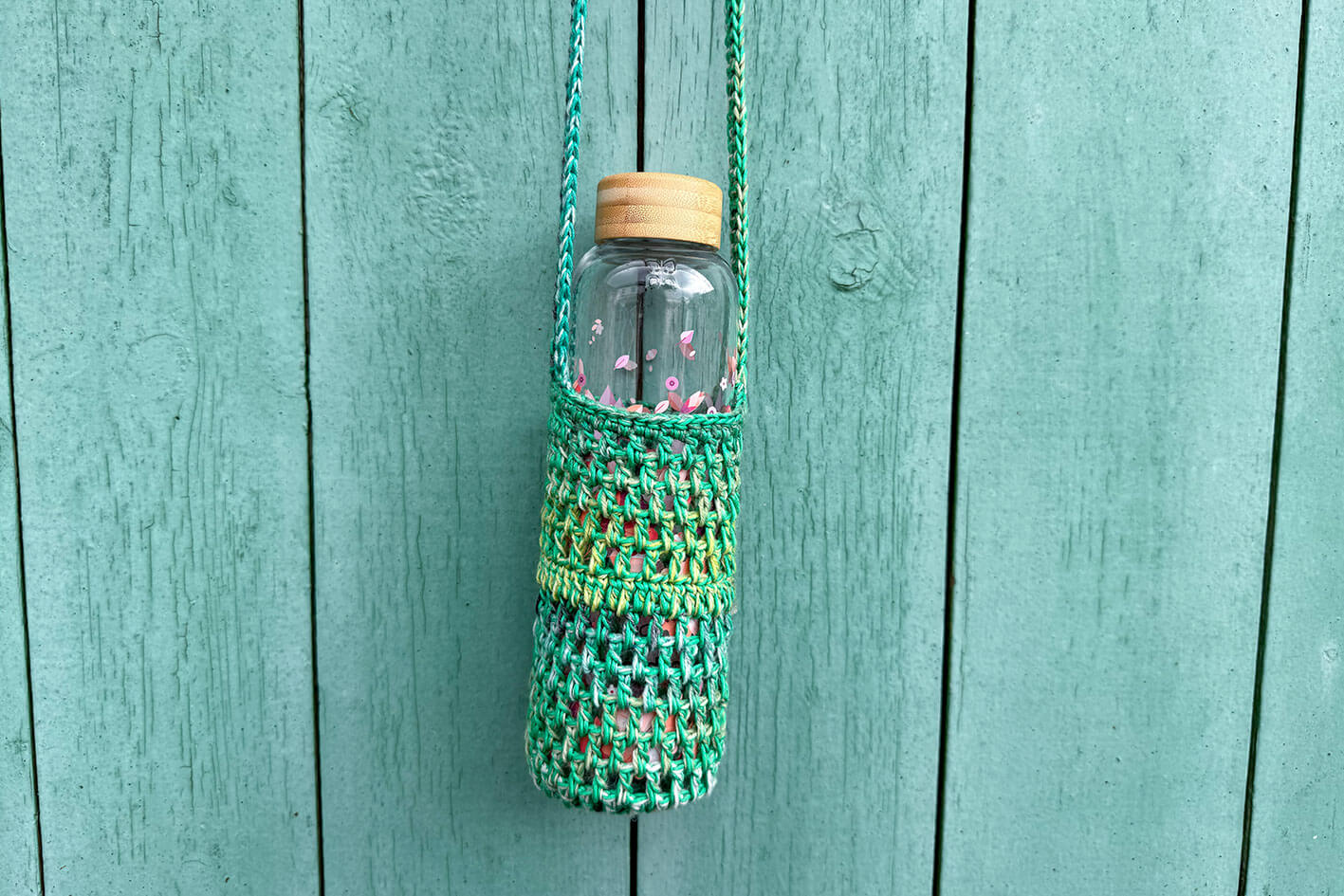 Gehäkelter, Flaschenhalter CHRISTA von Jeff Every Piece A Friend vor blauem Hintergrund. Der Flaschenhalter ist aus Baumwolle und kommt in verschiedenen Grüntönen. 