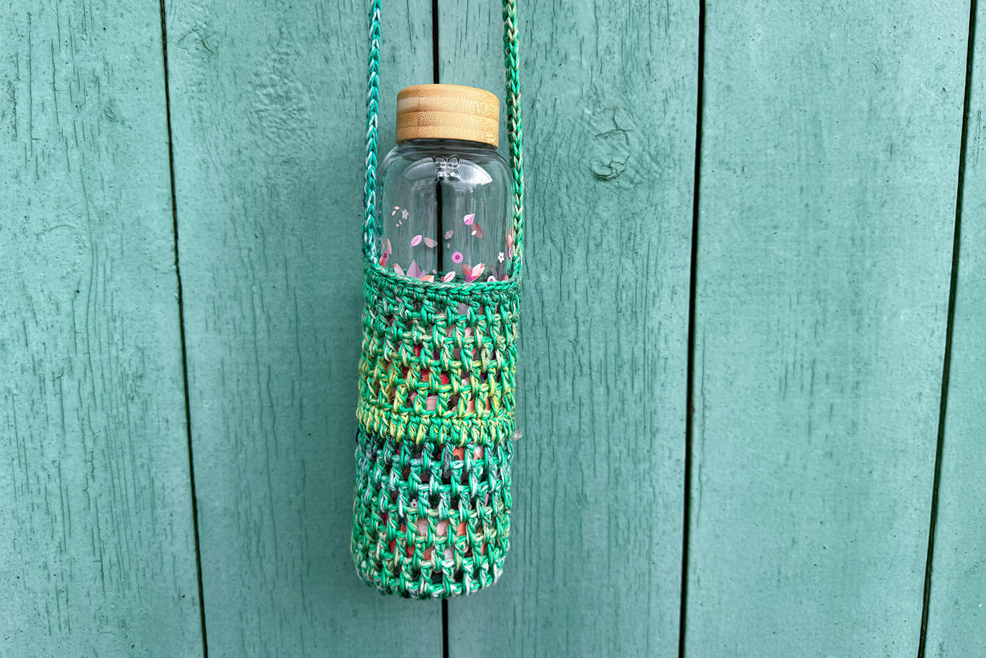Gehäkelter, Flaschenhalter CHRISTA von Jeff Every Piece A Friend vor blauem Hintergrund. Der Flaschenhalter ist aus Baumwolle und kommt in verschiedenen Grüntönen. 