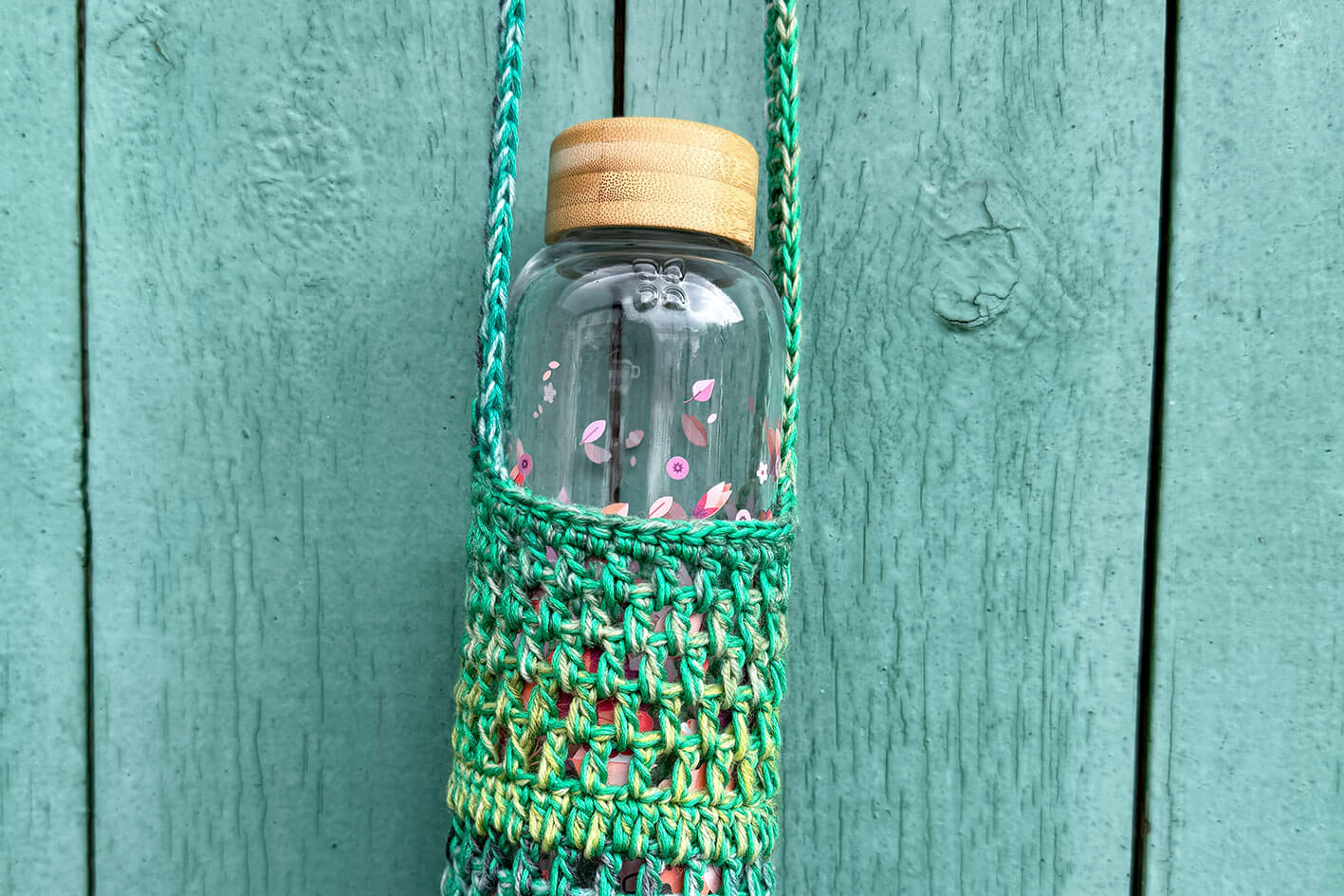Detailansicht: Gehäkelter, Flaschenhalter CHRISTA von Jeff Every Piece A Friend vor blauem Hintergrund. Der Flaschenhalter ist aus Baumwolle und kommt in verschiedenen Grüntönen. 