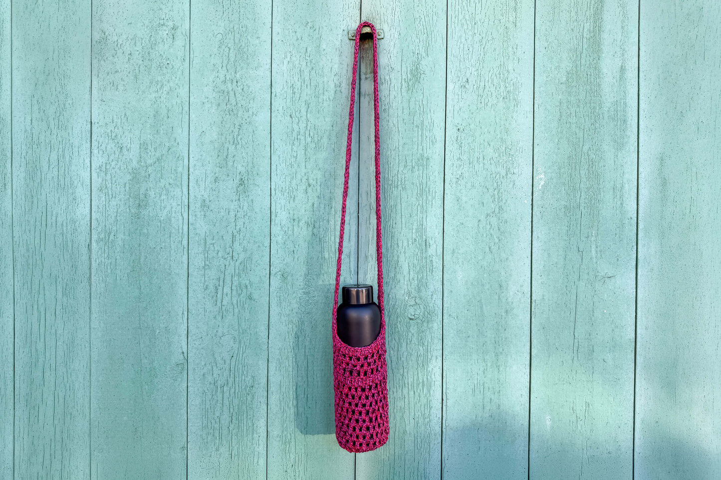 Gehäkelter, Flaschenhalter CHRISTA von Jeff Every Piece A Friend vor blauem Hintergrund. Der Flaschenhalter hat die Farbe pink oder magenta gemischt.