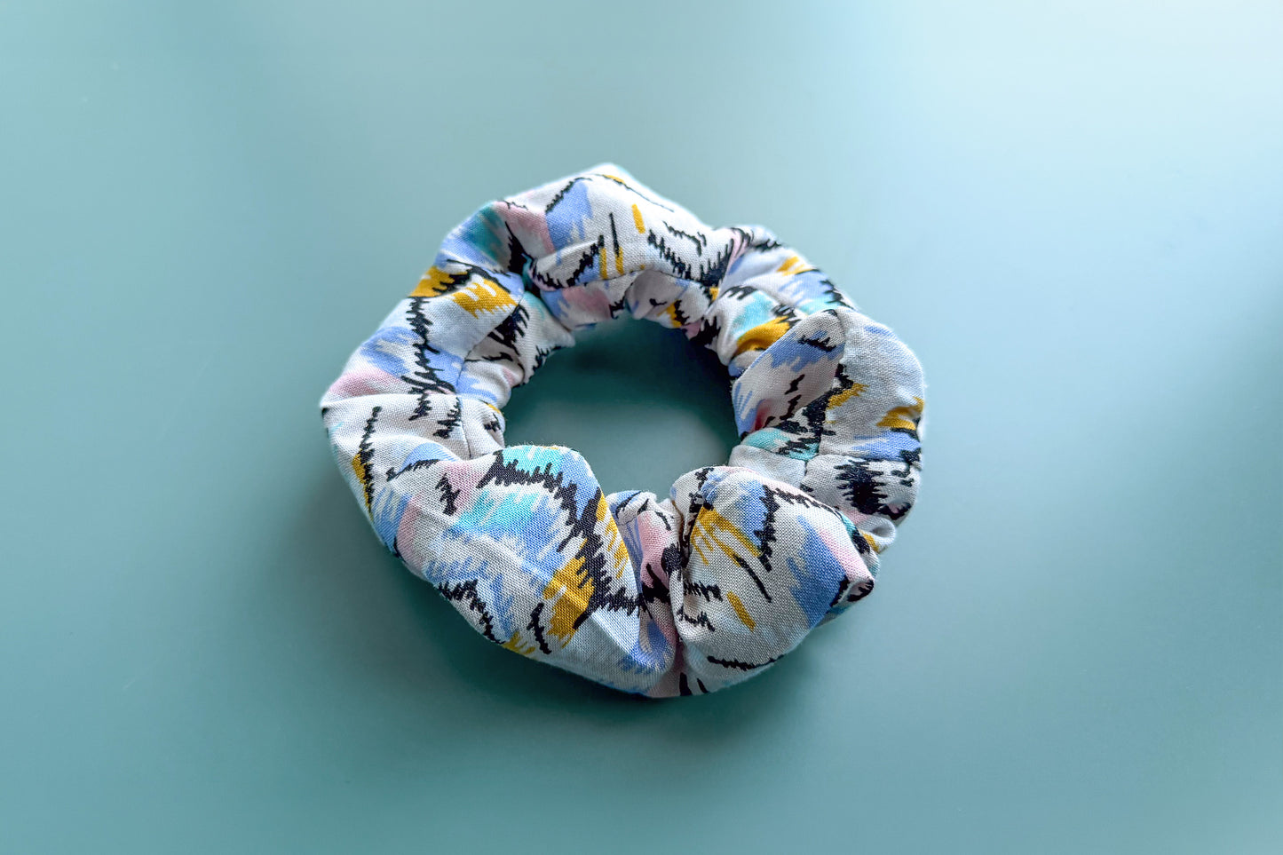 Ansicht von schräg oben: Ein beiger Scrunchie mit buntem Muster und Reißverschluss liegt auf einem blauen Tisch. Der Reißverschluss ist geschlossen.  Jeff - EveryPieceAFriend