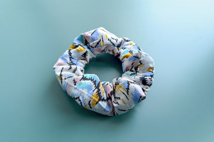 Ansicht von schräg oben: Ein beiger Scrunchie mit buntem Muster und Reißverschluss liegt auf einem blauen Tisch. Der Reißverschluss ist geschlossen.  Jeff - EveryPieceAFriend