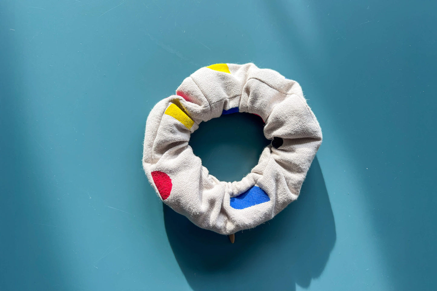 Ansicht von oben: ein beiger Scrunchie mit beigen Reißverschluss, gemustert mit geometrischen, roten, blauen und gelben Flächen liegt auf einem blauen Tisch. Der Reißverschluss ist geschlossen.