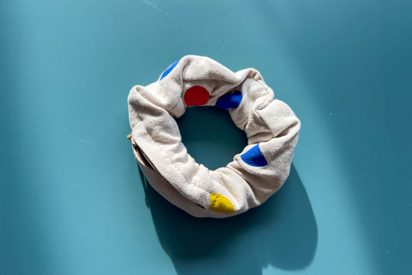 Ansicht von oben: ein beiger Scrunchie mit beigen Reißverschluss, gemustert mit geometrischen, roten, blauen und gelben Flächen liegt auf einem blauen Tisch. Der Reißverschluss ist offen.