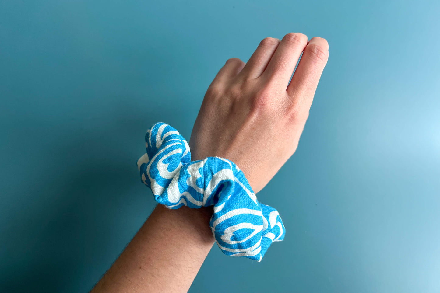 Eine Hand trägt einen hellblau-weißen Scrunchie am Handgelenk vor blauem Hintergrund. Der Scrunchie besteht aus upcycled Materialien und hat einen Reißverschluss.