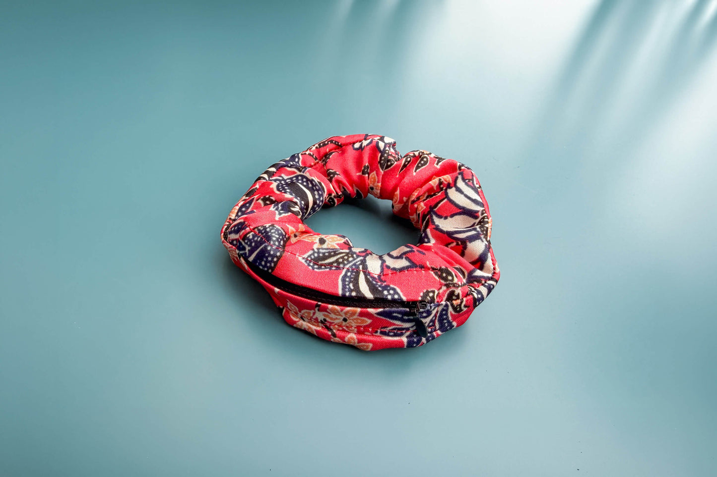Ansicht von oben: Ein altrosa Scrunchie mit Reißverschluss und weiß-schwarz-orangem Blumenmuster liegt auf einem blauen Tisch. Der Reißverschluss ist geschlossen.  Jeff - everyPieceAFriend