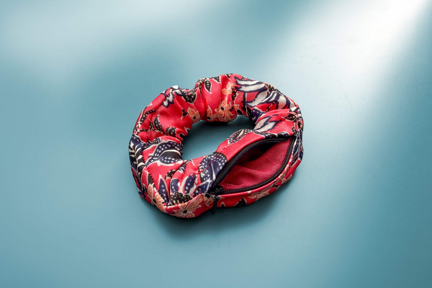 Ansicht von oben: Ein altrosa Scrunchie mit Reißverschluss und weiß-schwarz-orangem Blumenmuster liegt auf einem blauen Tisch. Der Reißverschluss ist geöffnet.  Jeff - everyPieceAFriend
