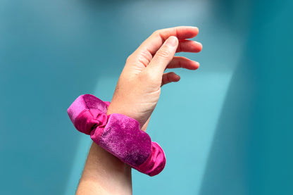 Eine Hand trägt einen magenta-lila Scrunchie am Handgelenk vor blauem Hintergrund. Der Scrunchie besteht aus upcycled Materialien, lila Samt- und magenta Baumwollstoff und hat einen Reißverschluss.