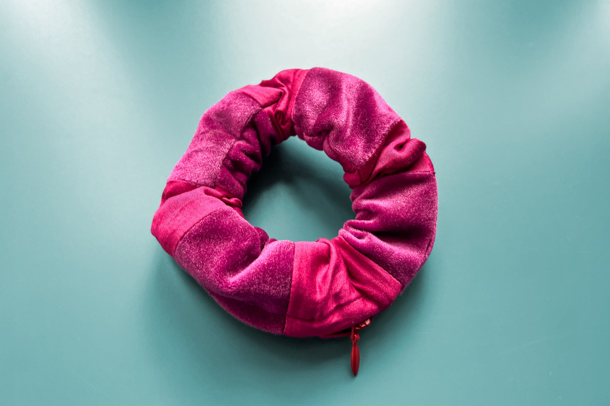 Ansicht von oben: Ein magenta-lila Scrunchie mit Reißverschluss und liegt auf einem blauen Tisch. Der Schrunchie besteht aus magenta Baumwoll- und lila Samstoff. Er hat einen Reißverschluss, dieser ist Reißverschluss geschlossen.