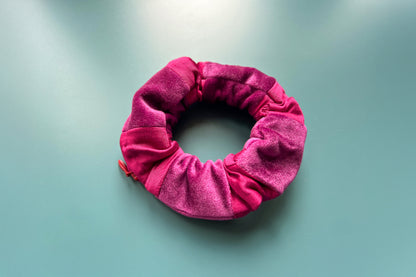 Ansicht von leicht schräg oben: Ein magenta-lila Scrunchie mit Reißverschluss und liegt auf einem blauen Tisch. Der Schrunchie besteht aus magenta Baumwoll- und lila Samstoff. Er hat einen Reißverschluss, dieser ist Reißverschluss geschlossen.
