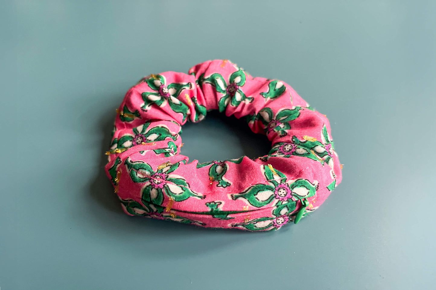 Ansicht von oben: Ein Scrunchie mit Reißverschluss und einem Blumenmuster in roten und grünen Tönen liegt auf einem blauen Tisch. Der Reißverschluss ist geschlossen.