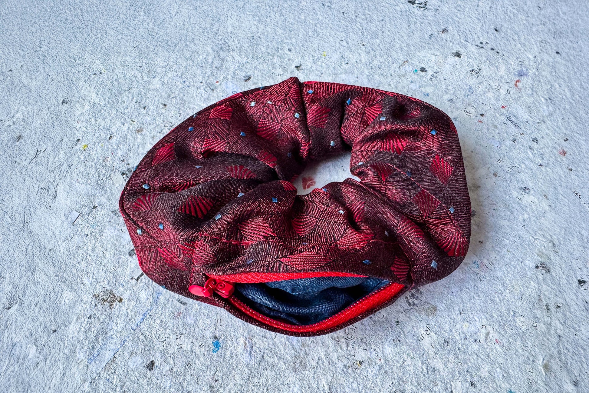 Ansicht von oben: Ein Scrunchie mit Reißverschluss und bordeauxfarbenen Muster liegt auf einem grauem Untergrund. Der rote Reißverschluss ist geöffnet.