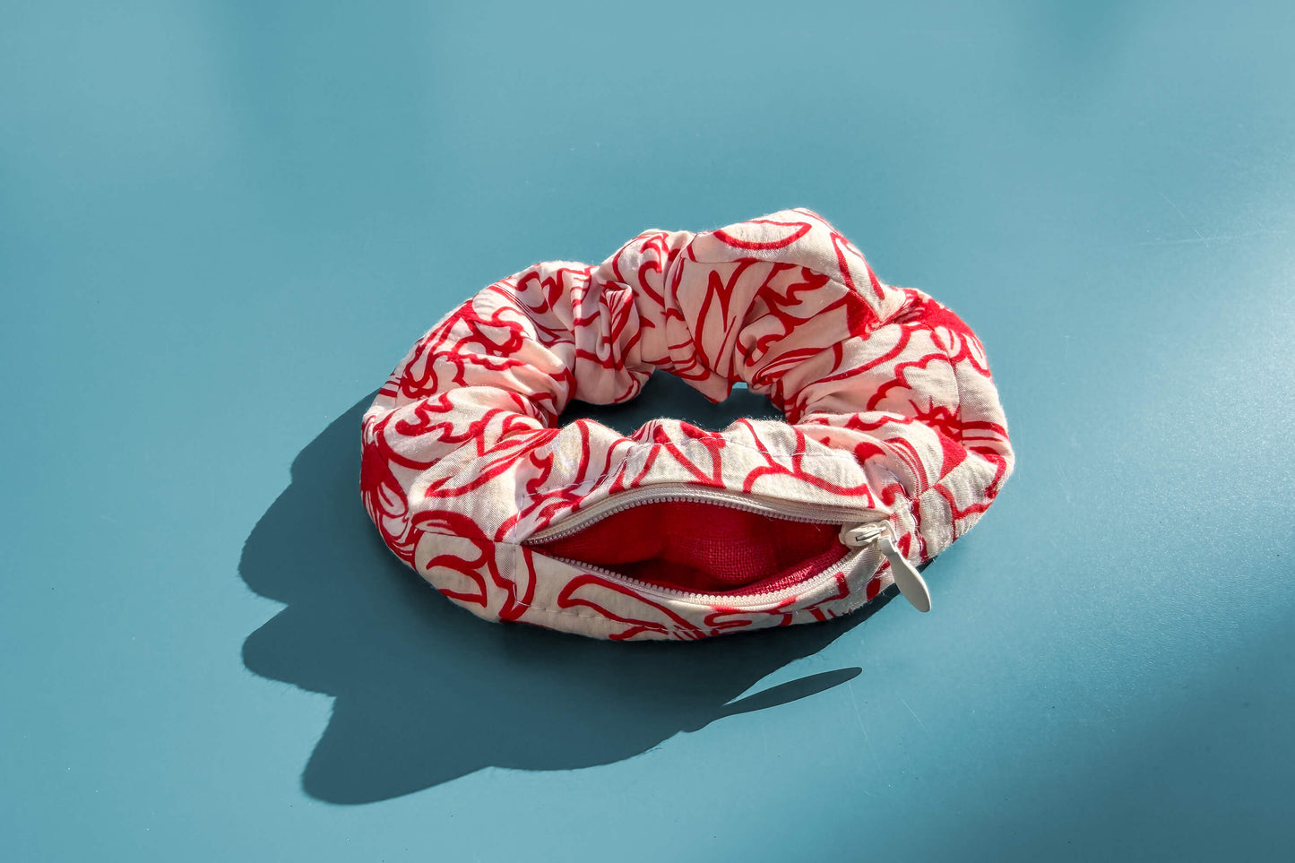 Ansicht von oben: Ein Scrunchie mit geöffnetem Reißverschluss und weiß-rotem Blumenmuster liegt auf einem blauen Tisch. Der Reißverschluss ist geöffnet. Jeff - everyPieceAFriend