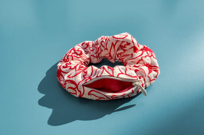 Ansicht von oben: Ein Scrunchie mit geöffnetem Reißverschluss und weiß-rotem Blumenmuster liegt auf einem blauen Tisch. Der Reißverschluss ist geöffnet. Jeff - everyPieceAFriend