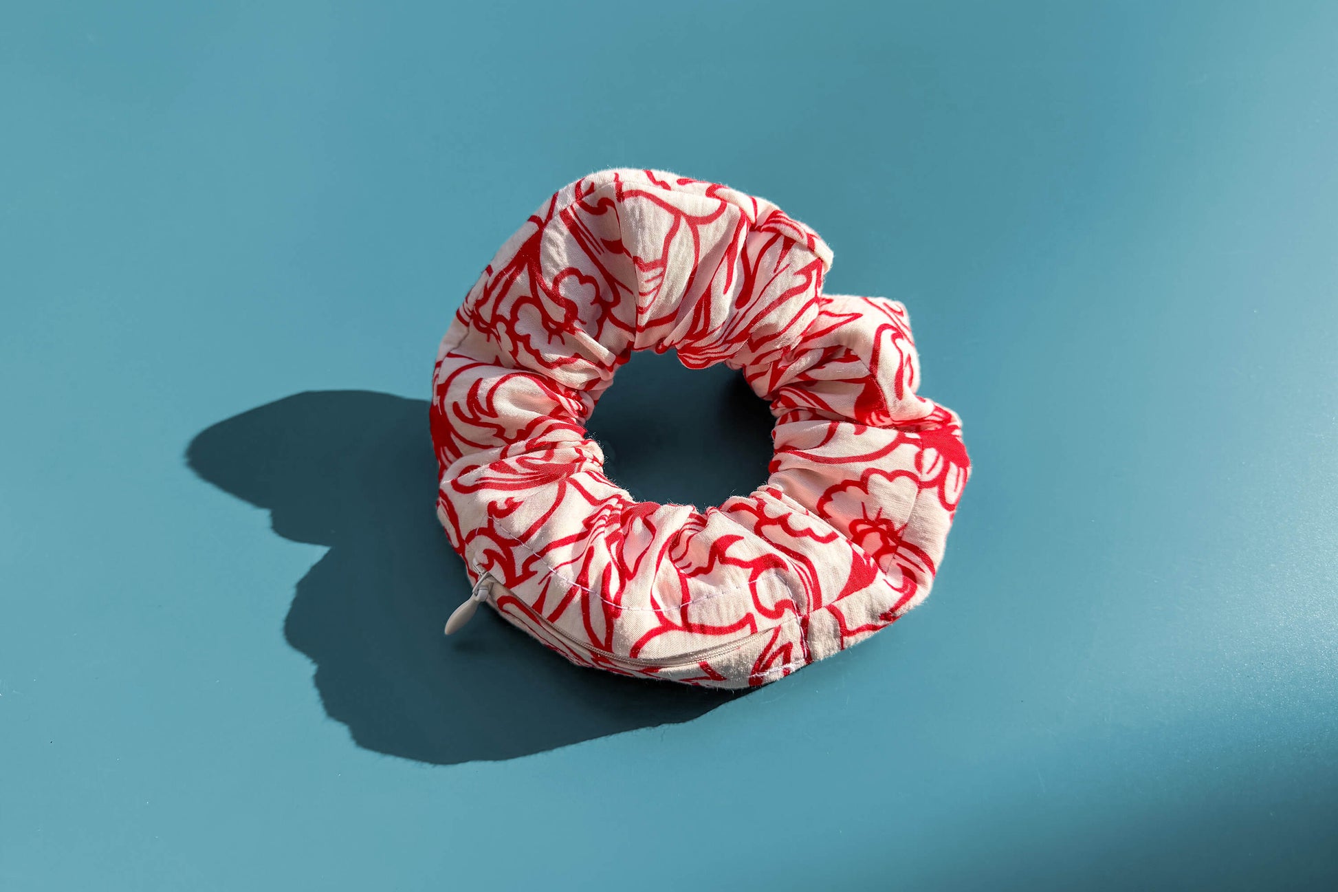 Ansicht von oben: Ein Scrunchie mit Reißverschluss und weiß-rotem Blumenmuster liegt auf einem blauen Tisch. Jeff - everyPieceAFriend