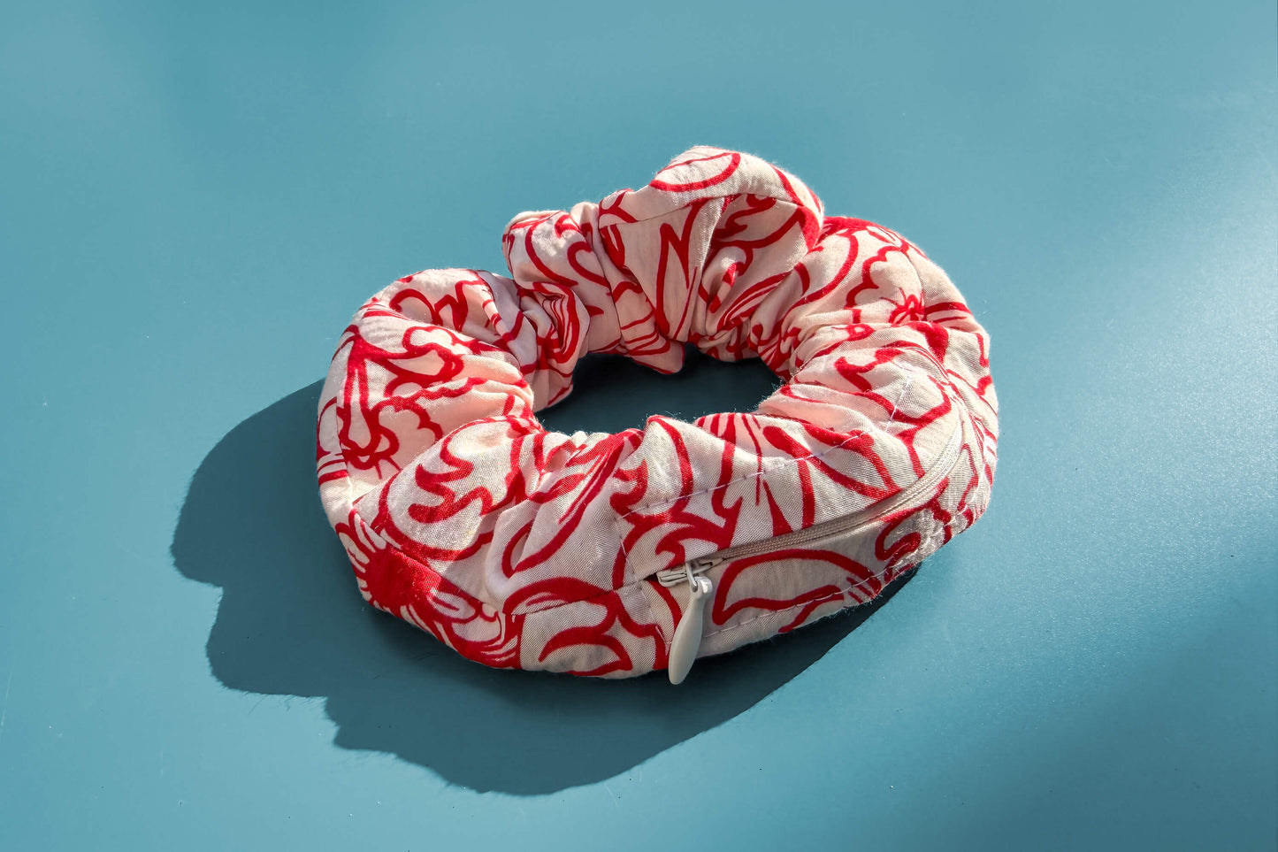 Ansicht von oben: Ein Scrunchie mit Reißverschluss und weiß-rotem Blumenmuster liegt auf einem blauen Tisch. Der Reißverschluss ist geöffnet.  Jeff - everyPieceAFriend