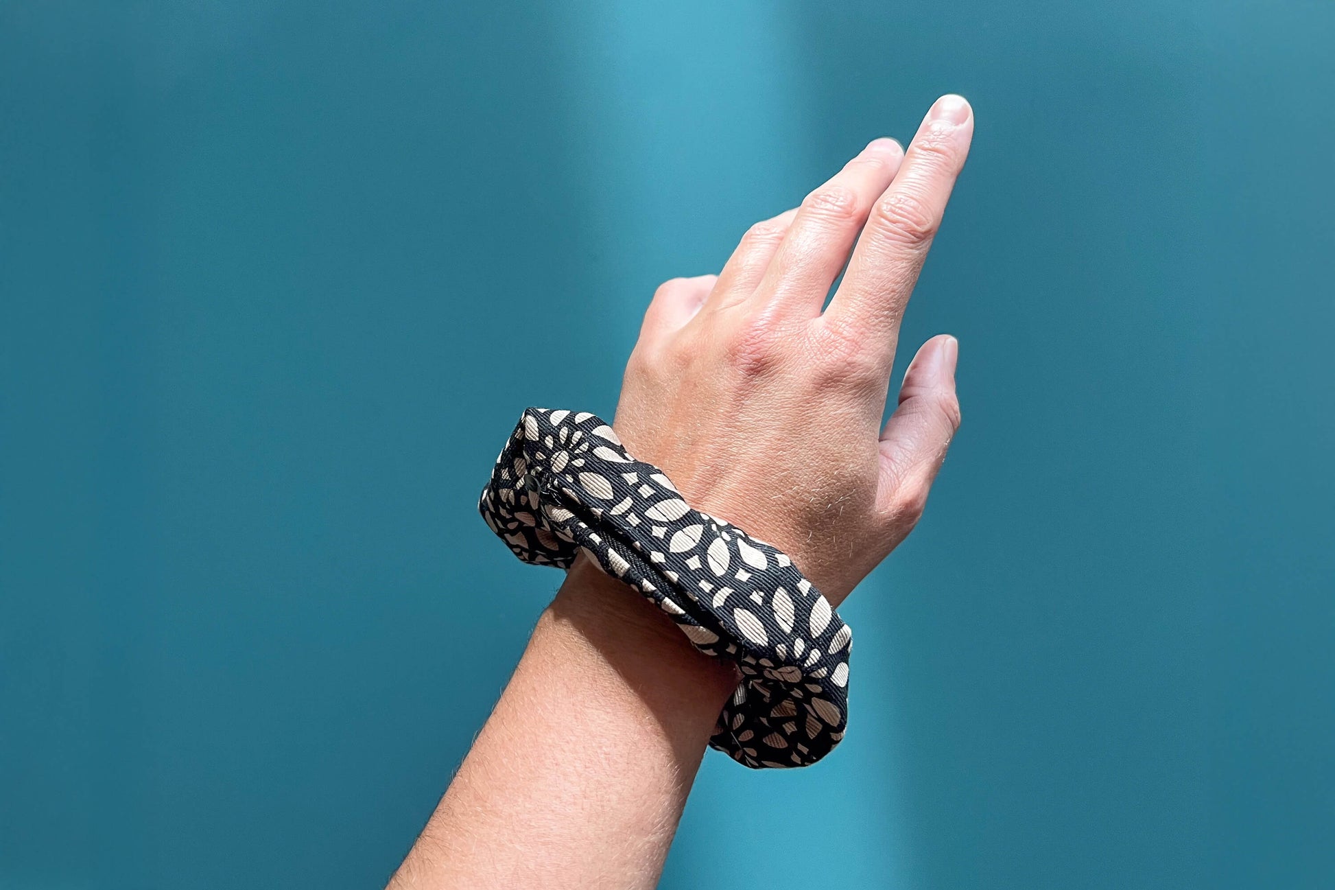 Eine Hand trägt einen schwarzen Scrunchie mit beigen Blumenmuster am Handgelenk vor blauem Hintergrund. Der Scrunchie besteht aus upcycled Materialien und hat einen schwarzen Reißverschluss.