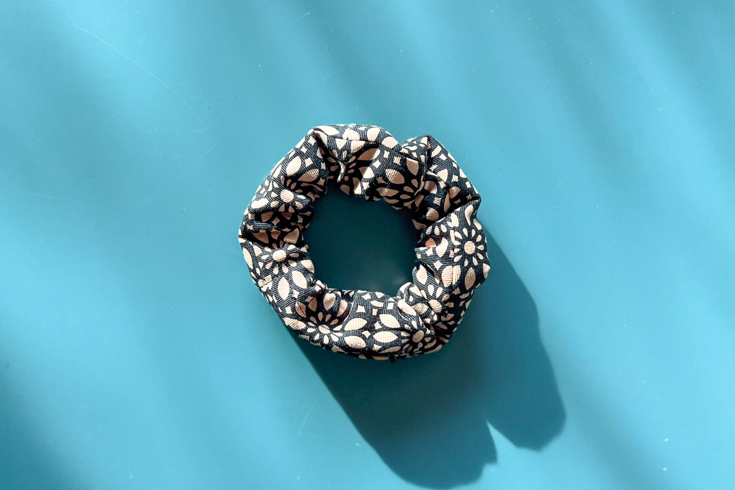 Ansicht von oben: Ein Scrunchie mit Reißverschluss und schwarz-beigen Blumenmuster liegt auf einem blauen Tisch. Der Reißverschluss ist geschlossen.