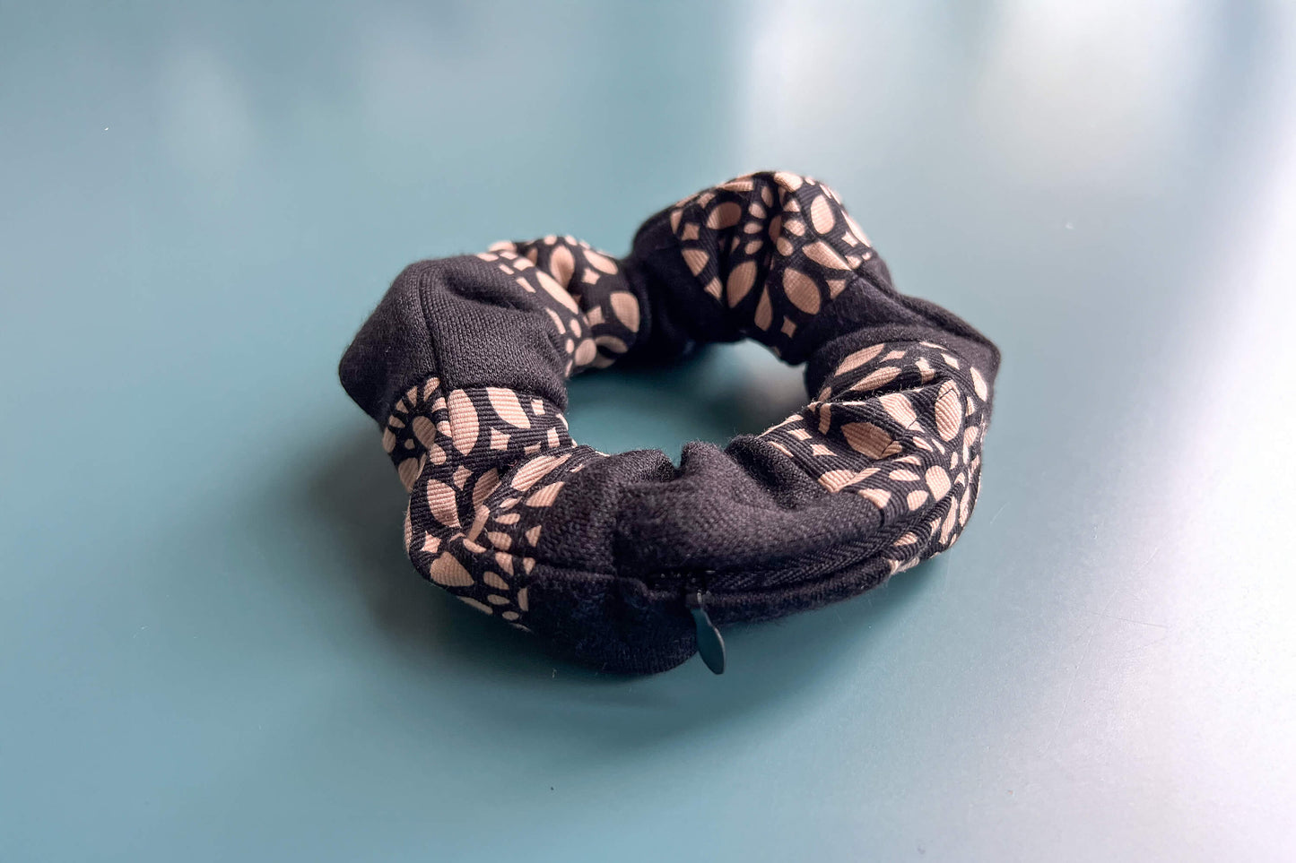 Ansicht von schräg oben: Ein schwarz-beige gestreifter Scrunchie mit schwarzem Reißverschluss und schwarz-beigen Blumenmuster liegt auf einem blauen Tisch. Der Reißverschluss ist geschlossen.