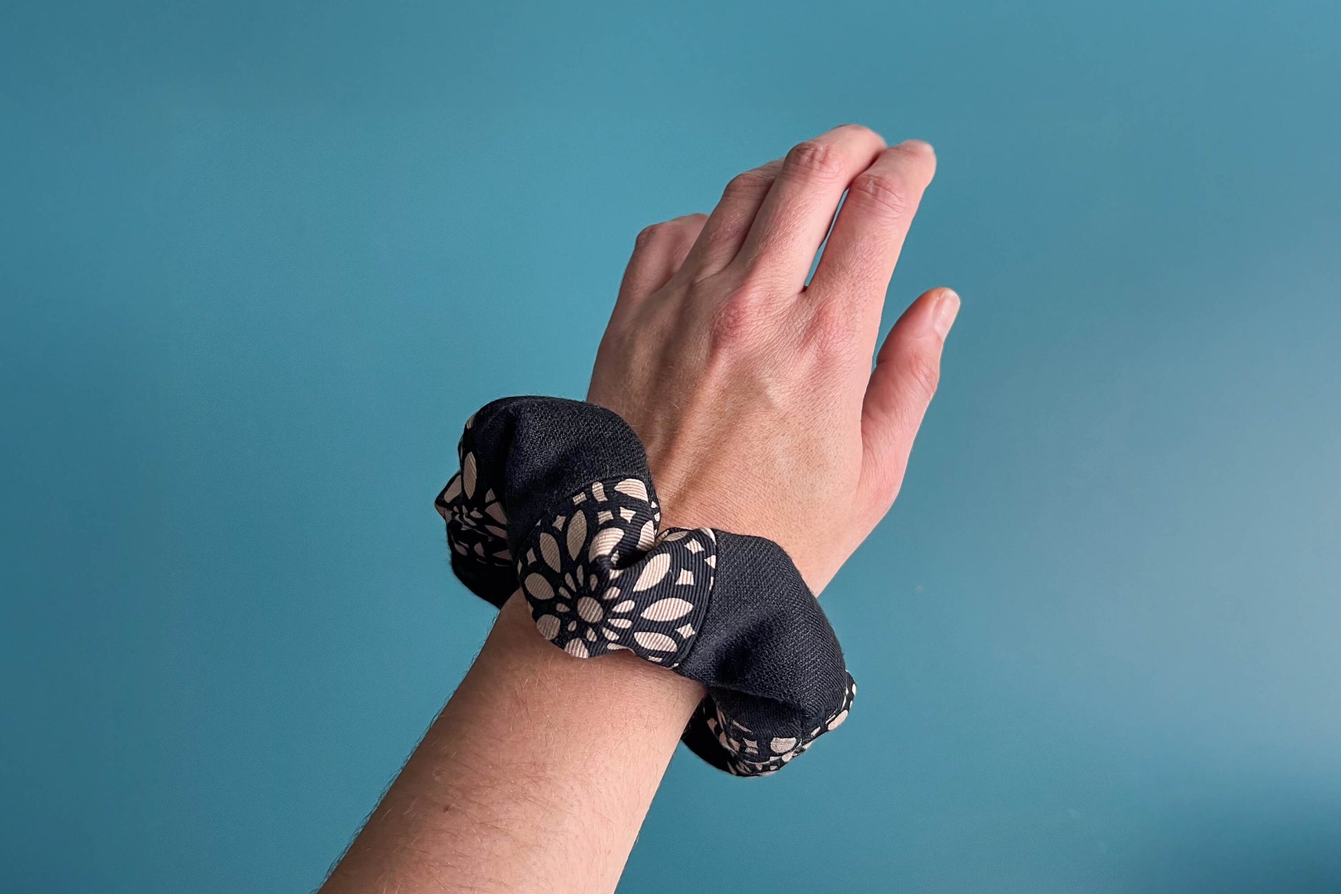Eine Hand trägt einen schwarz-beige gestreiften Scrunchie mit schwarzem Reißverschluss und schwarz-beigen Blumenmuster am Handgelenk. Der Reißverschluss ist geschlossen. Blauer Hintergrund