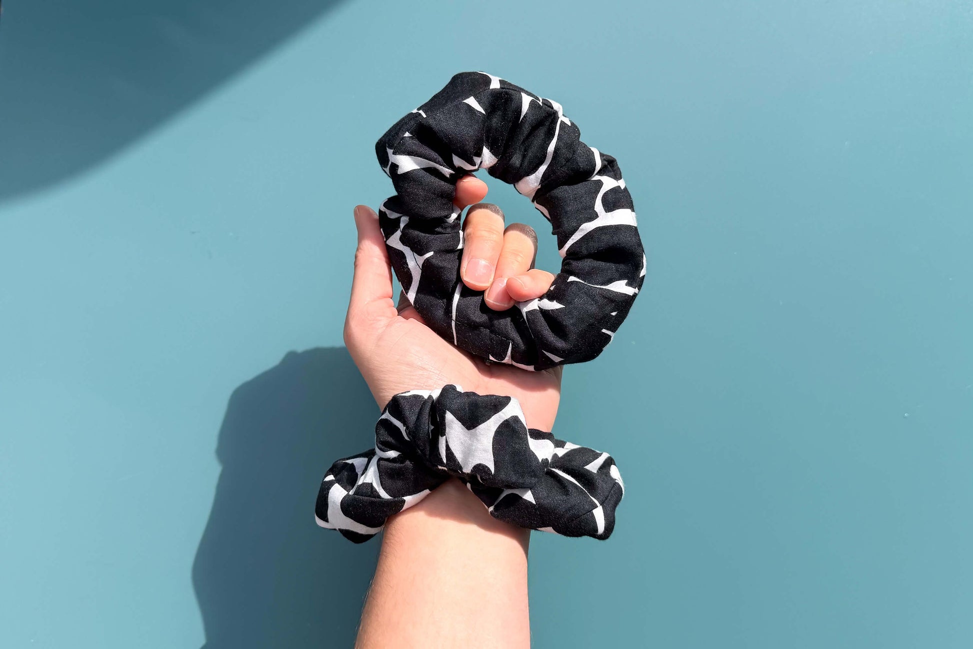 Ansicht von oben: Ein Hand trägt zwei schwarz-weiße Scrunchies mit schwarzen Reißverschluss. Blauer Hintergrund. Der Reißverschluss ist geschlossen.  Jeff - EveryPieceAFriend