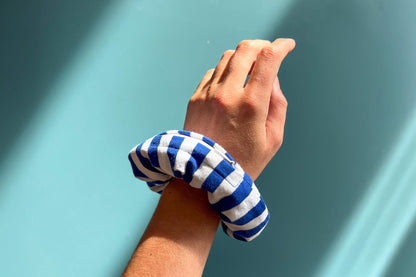 Eine Hand trägt einen dunkelblau-weißen gestreiften Scrunchie am Handgelenk vor blauem Hintergrund. Der Scrunchie besteht aus upcycled Materialien und hat einen weißen Reißverschluss.