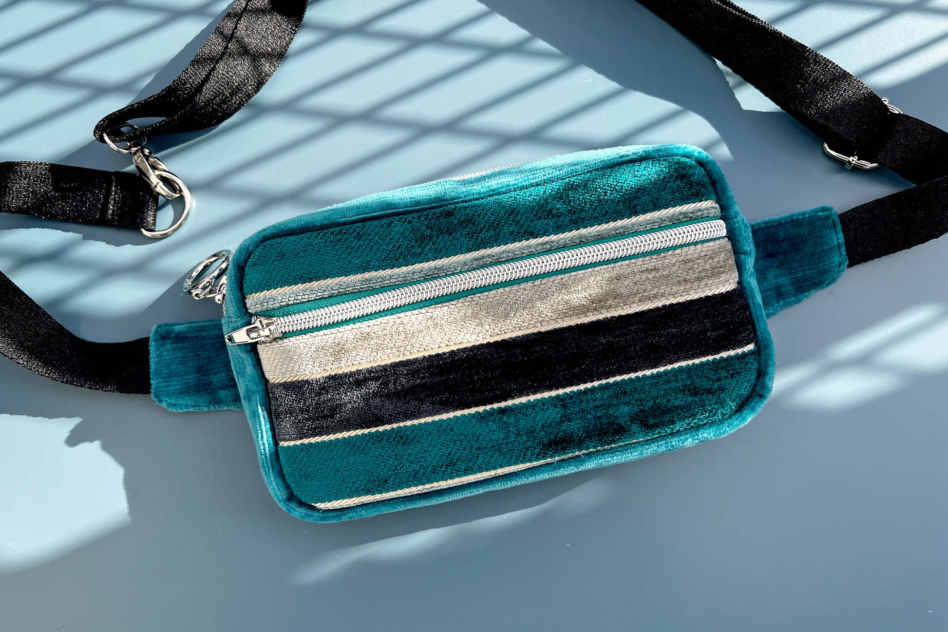 Gesamtansicht einer Bauchtasche aus Samt von Jeff EveryPieceAFriend. Die Tasche ist blau und grau gestreift, hat ein schwarzes Gurtband und einen silbernen Reißverschluss. 