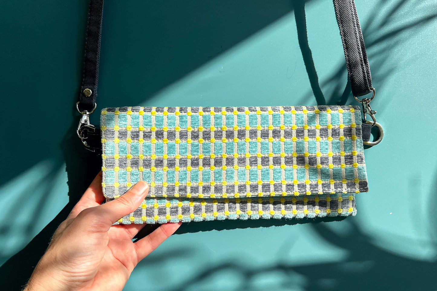 Gesamtansicht: Hand hält die Umhängetasche DANA aus upcycled Polsterstoff in den Farben blau, gelb und beige mit silbernen Verschlüssen in die Kamera. 