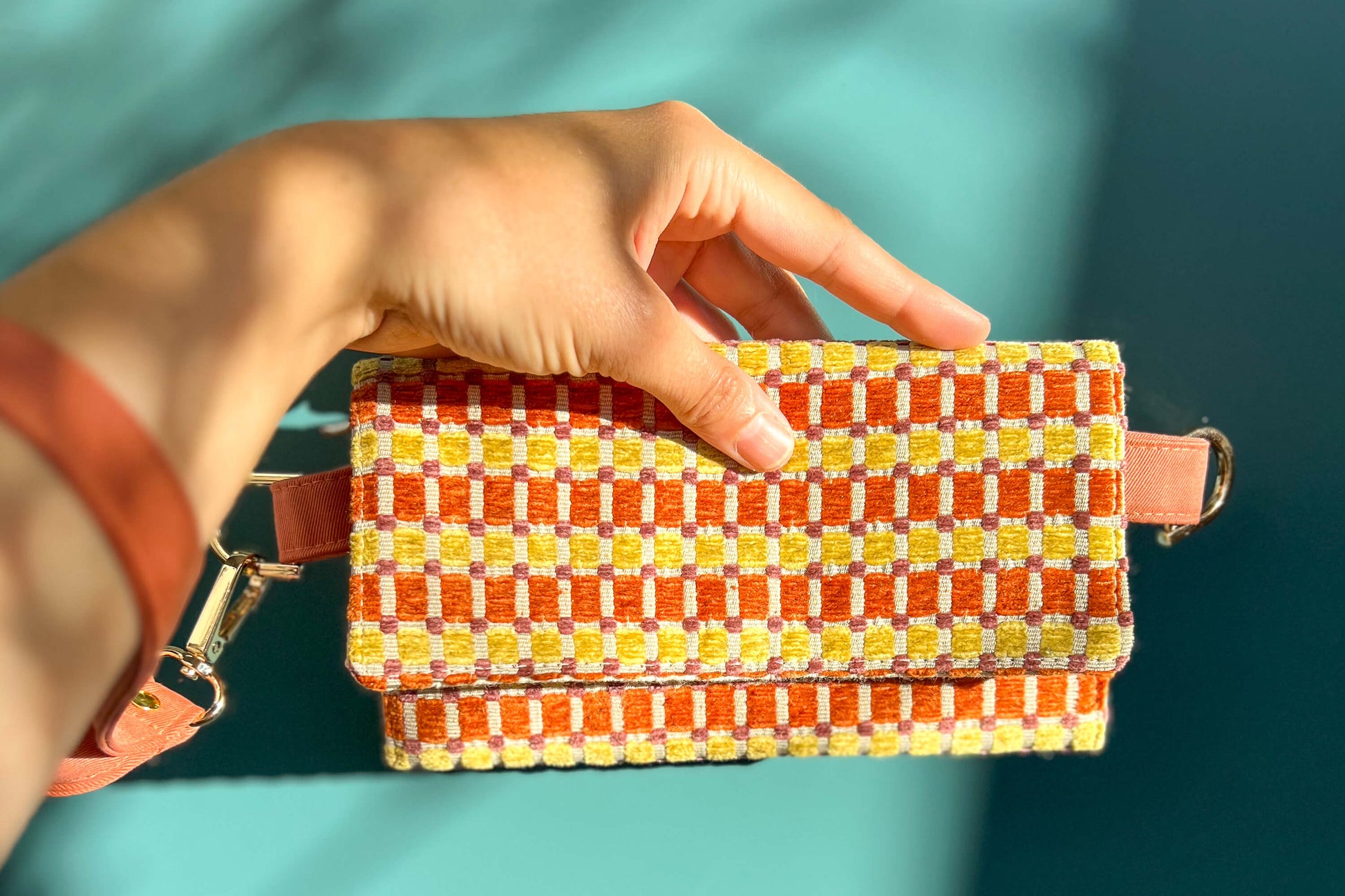 Gesamtansicht: eine Hand hält die Handtasche DANA in der Farbe gelb-orange-beige vor einem blauen Hintergrund ins Bild. 