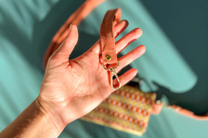 Eine Hand hält das Gurtband Handtasche DANA in der Farbe gelb-orange-beige vor einem blauen Hintergrund ins Bild.