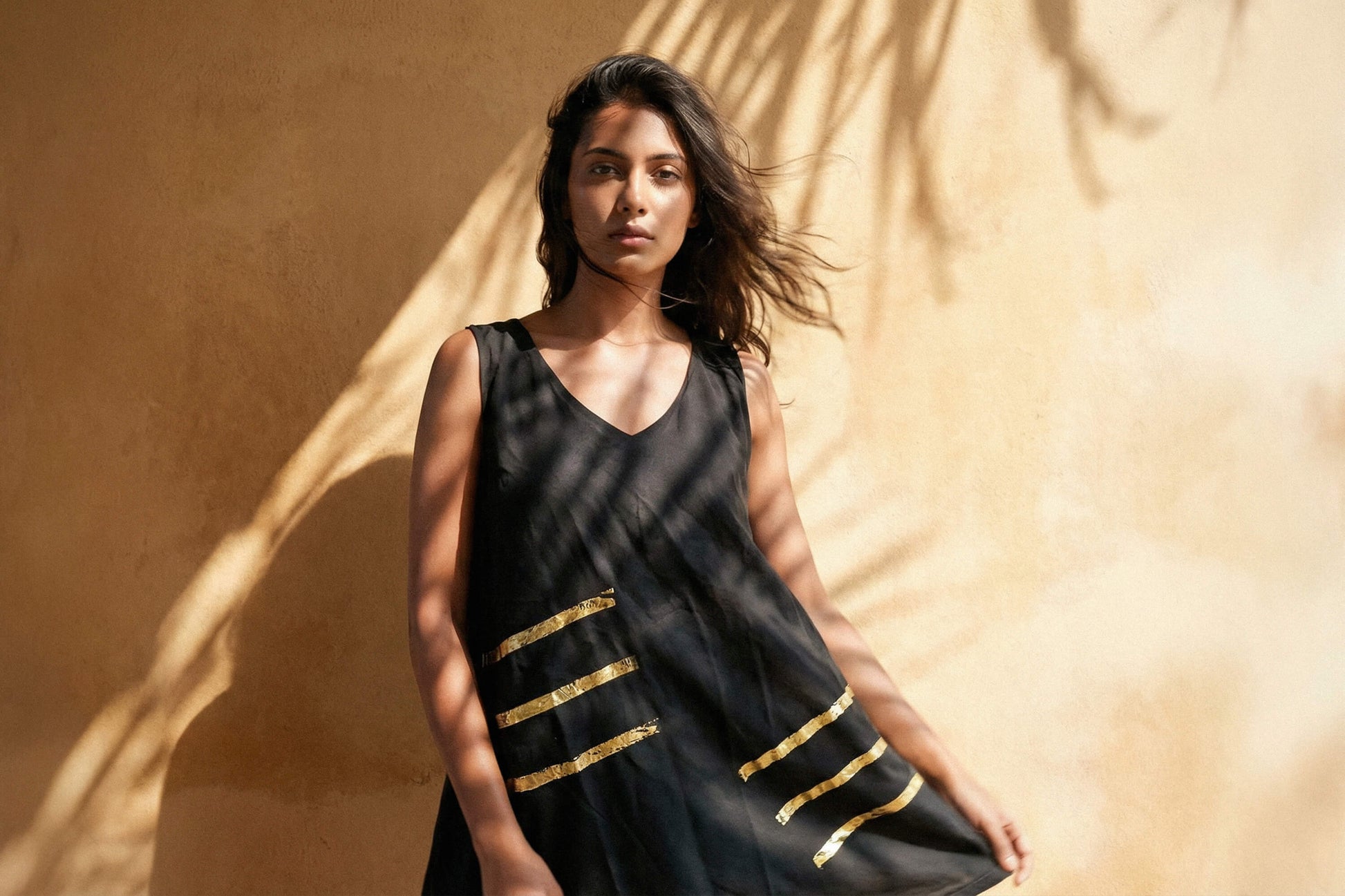 Eine junge Frau steht vor einer beigen Wand, sie sieht in die Kamera. Auf der Wand ist ein Licht- und Schattenspiel zu sehen. Sie trägt ein schwarzes Kleid mit einem goldenen, minimalistischen Muster. Das Kleid hat einen weiten Schnitt und endet über den Knien der Frau.