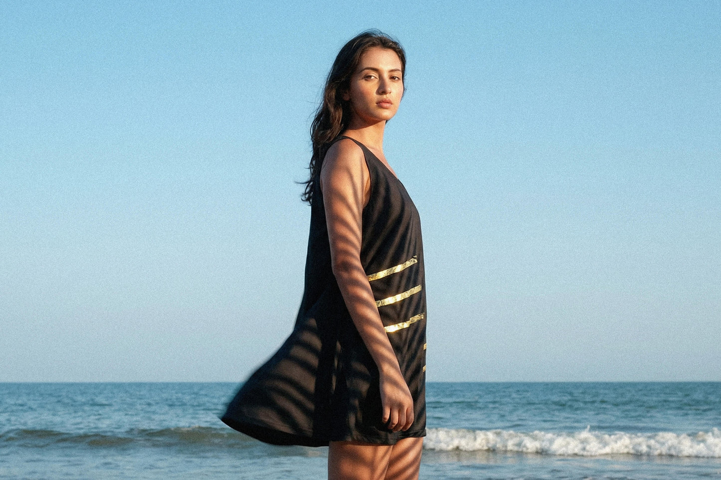 Junge Frau steht am Strand, wir sehen sie von der Seite, sie sieht in die Kamera. Im Hintergrund ist das Meer zu sehen. Sie trägt ein schwarzes Kleid mit einem goldenen, minimalistischen Muster. Das Kleid hat einen weiten Schnitt und endet über den Knien der Frau.
