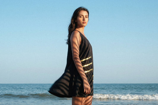 Junge Frau steht am Strand, wir sehen sie von der Seite, sie sieht in die Kamera. Im Hintergrund ist das Meer zu sehen. Sie trägt ein schwarzes Kleid mit einem goldenen, minimalistischen Muster. Das Kleid hat einen weiten Schnitt und endet über den Knien der Frau.