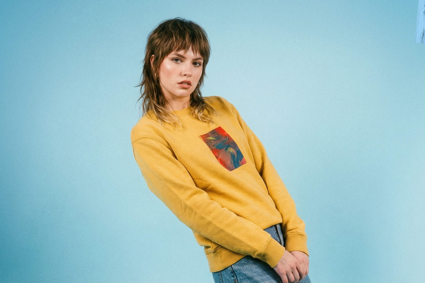 Junge Frau trägt senffarbenes Unisex Sweatshirt mit rot-blau-gelb verlaufendem Druck von einem großen Rechteck auf der Brust, bedruckt mit Siebdruck.