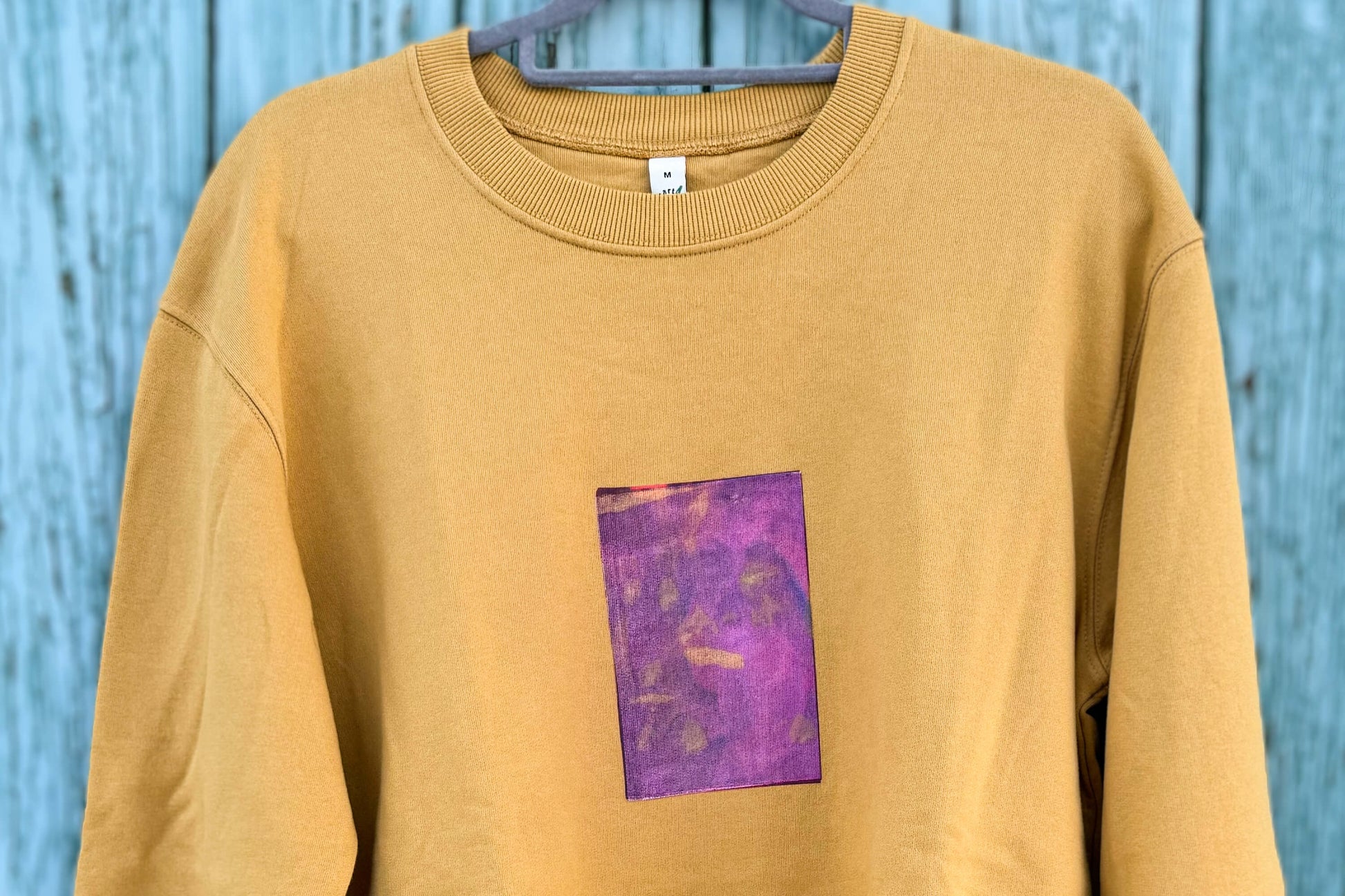 Detailansicht von senffarbenem senffarbenes Unisex Sweatshirt mit lila-gelb verlaufendem Druck von einem großen Rechteck auf der Brust, bedruckt mit Siebdruck. Siebdruck von Jeff EveryPieceAFriend.