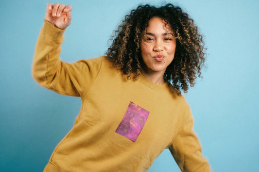 Junge Frau trägt senffarbenes Unisex Sweatshirt mit lila-gelb verlaufendem Druck von einem großen Rechteck auf der Brust, bedruckt mit Siebdruck.