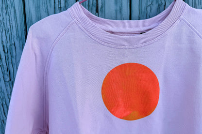 Detailansicht von light pink Unisex Sweatshirt mit orangenem, kreisrundem auf der Brust vor blauem Hintergrund, bedruckt mit Siebdruckfarben auf Wasserbasis. Siebdruck von Jeff EveryPieceAFriend.