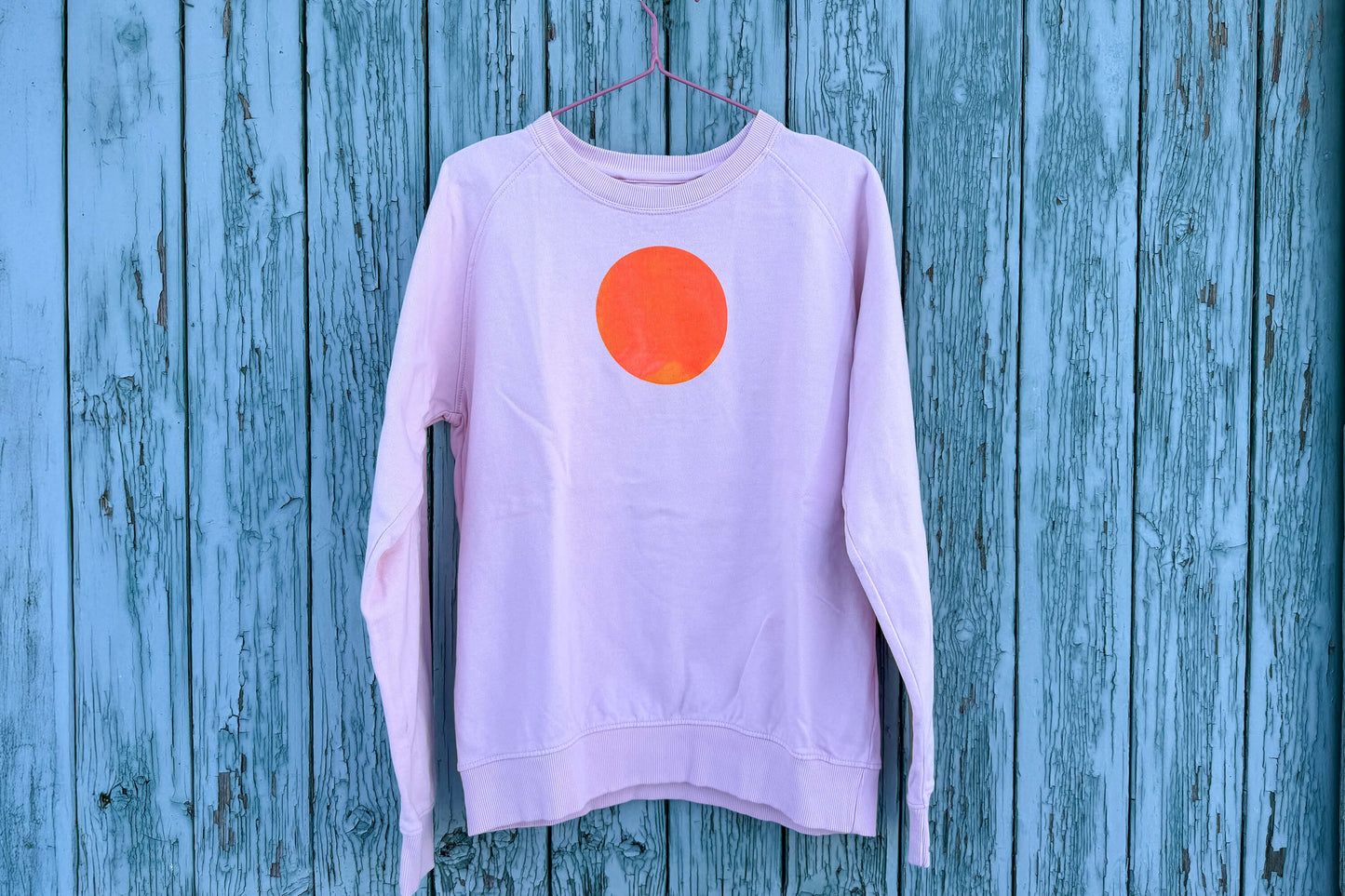 Gesamtansicht von light pink Unisex Sweatshirt mit orangenem, kreisrundem auf der Brust vor blauem Hintergrund, bedruckt mit Siebdruckfarben auf Wasserbasis. Siebdruck von Jeff EveryPieceAFriend.
