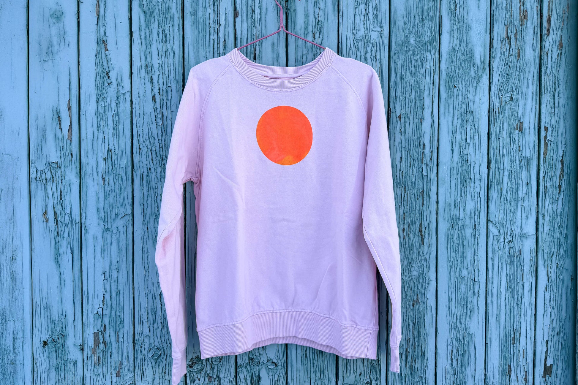 Gesamtansicht von light pink Unisex Sweatshirt mit orangenem, kreisrundem auf der Brust vor blauem Hintergrund, bedruckt mit Siebdruckfarben auf Wasserbasis. Siebdruck von Jeff EveryPieceAFriend.