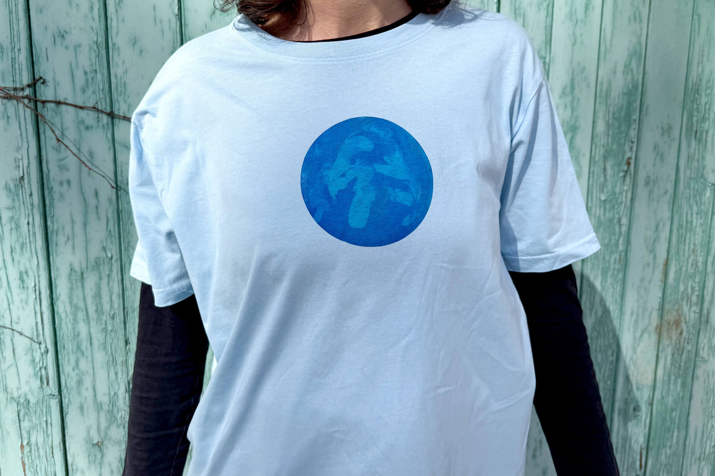 Frau trägt T-Shirt RONDA in hellblau mit einem rundem Aufdruck auf Brusthöhe. Das Shirt hängt zeigt einen Kreis auf Brusthöhe mit verschiedenen, dunklen Blautönen.