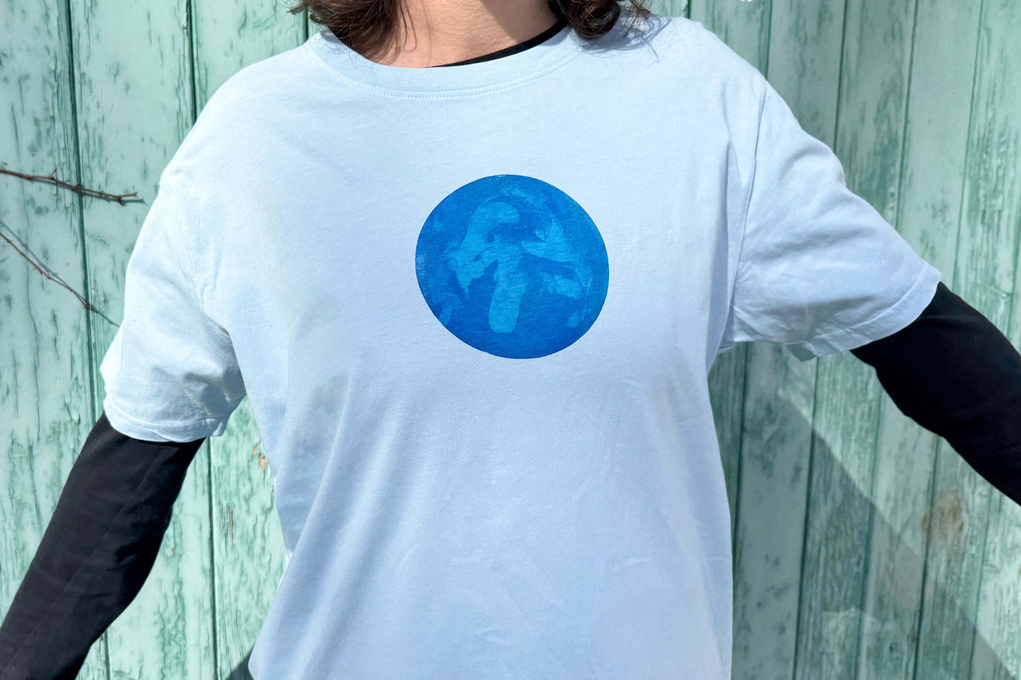 Frau trägt T-Shirt RONDA in hellblau mit einem rundem Aufdruck auf Brusthöhe. Das Shirt hängt zeigt einen Kreis auf Brusthöhe mit verschiedenen, dunklen Blautönen.