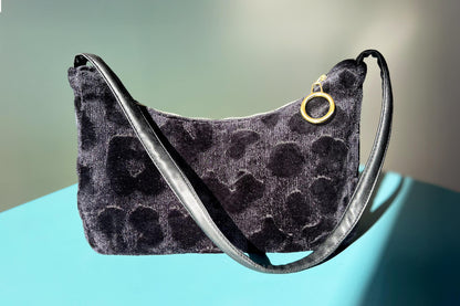 Gesamtansicht: Schultertasche im Vintagestil vor blau-grauem Hintergrund. Die Tasche ist schwarz gemustert und aus Samt und hat einen goldenen Reißverschluss. Die Tasche wurde aus upcycled Materialien von Jeff Every Piece A Friend hergestellt